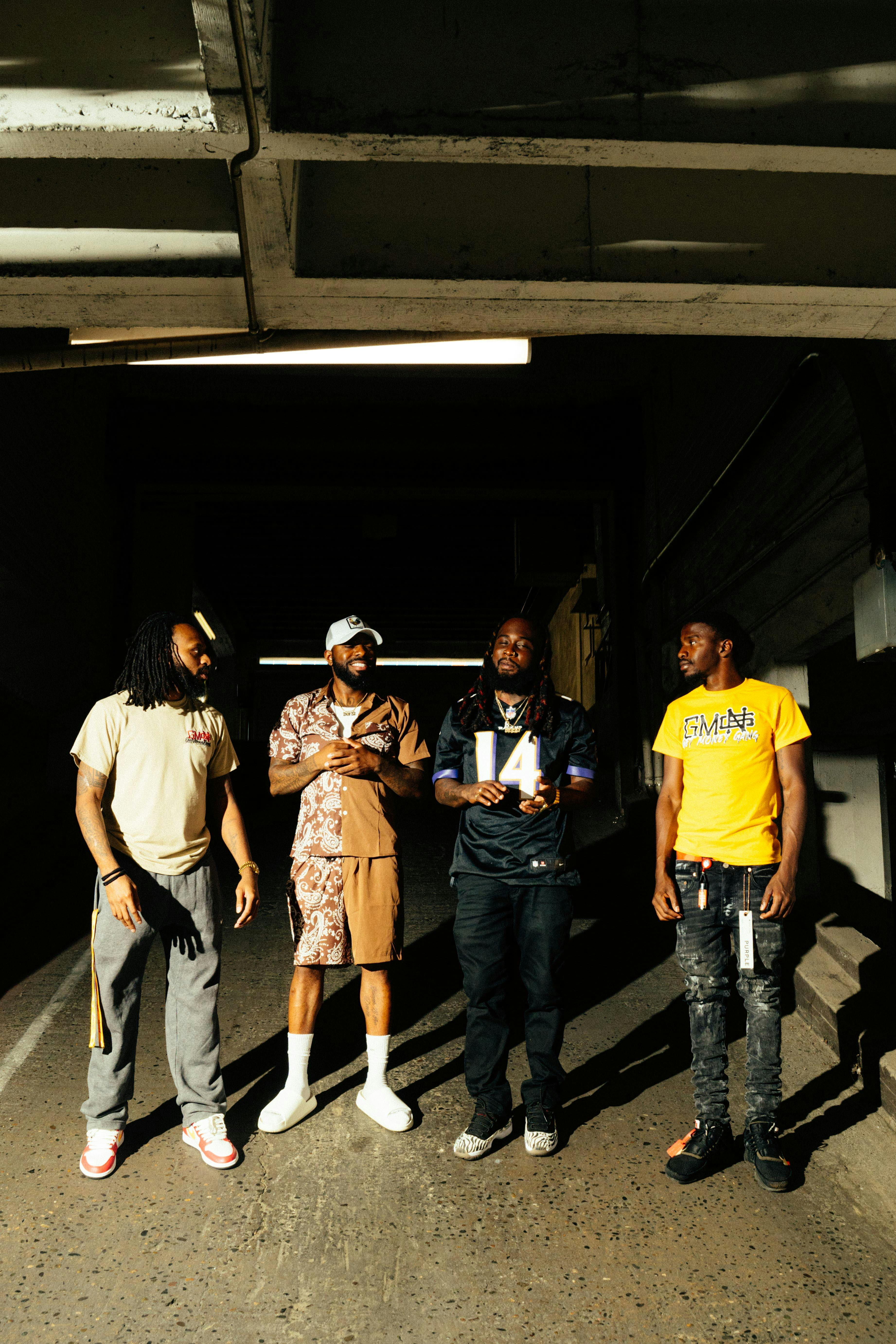 Four men standing together in sunlight under an overpass, showcasing urban fashion.