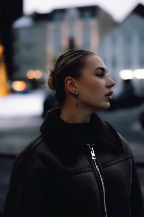 Free A stylish woman in a leather jacket stands on a city street at dusk, looking away. Stock Photo
