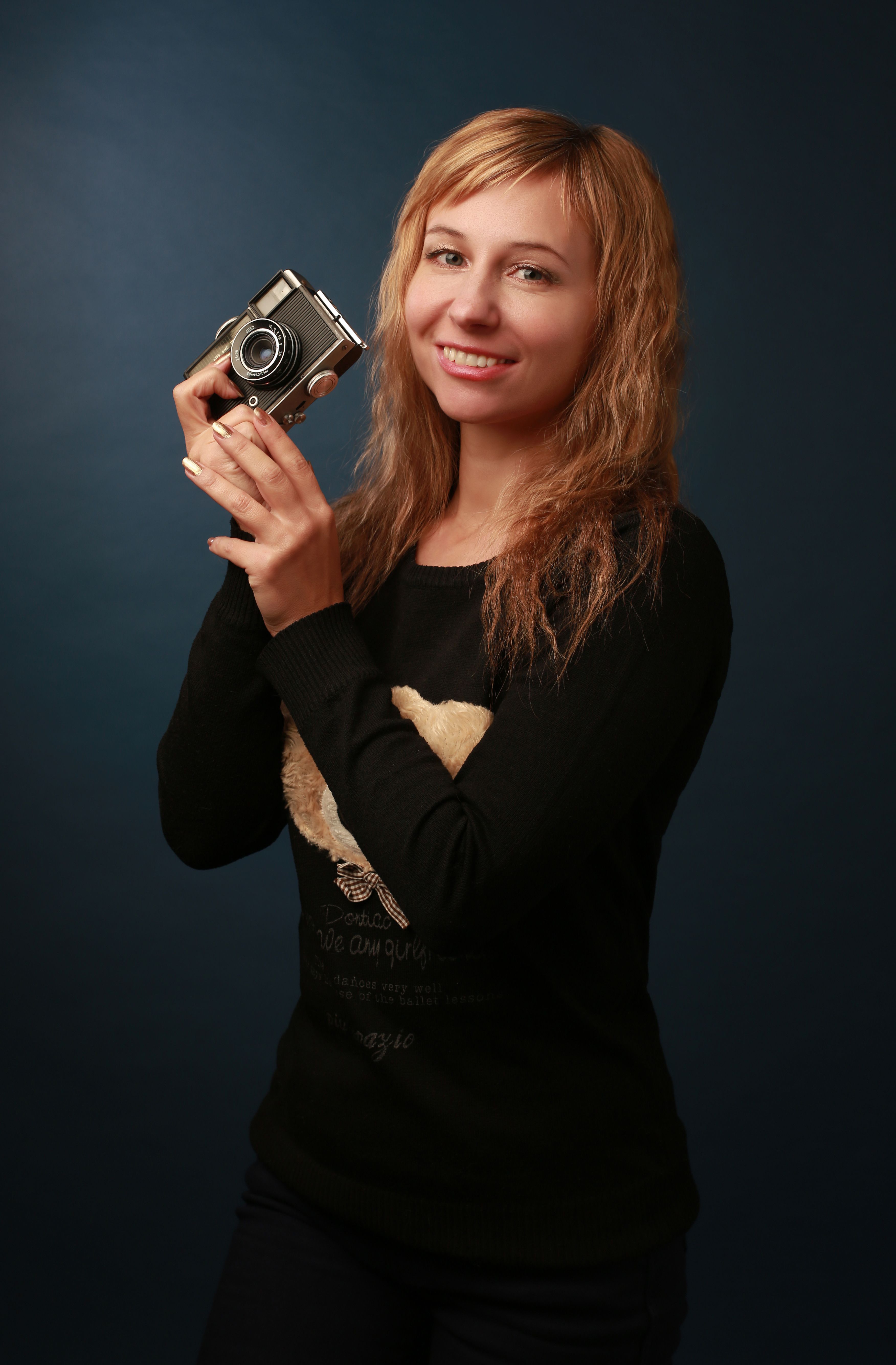 Smiling woman with long blonde hair holding a vintage camera against a dark backdrop.