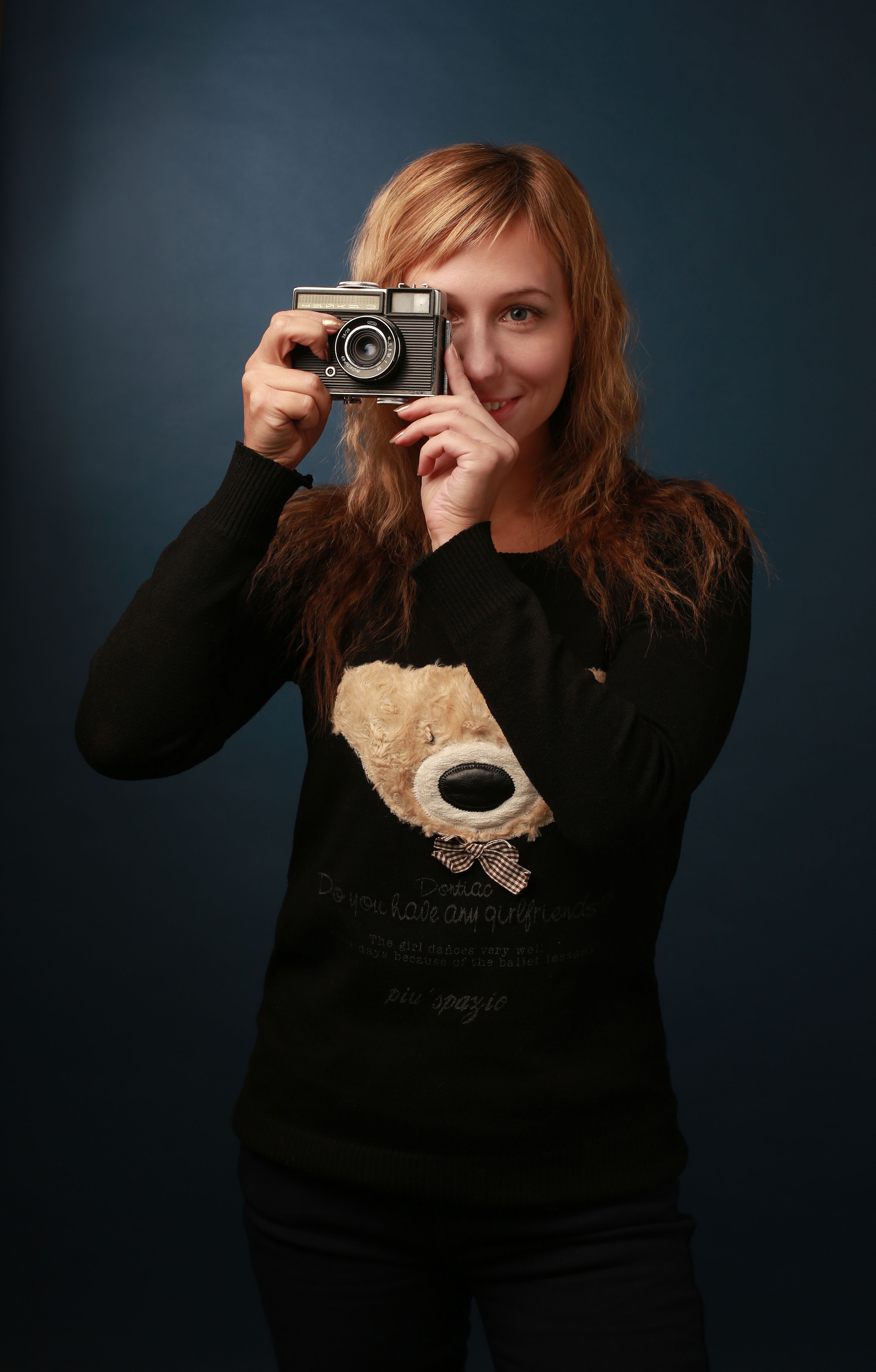 Blonde woman in black clothing holding a vintage camera, smiling for a portrait.