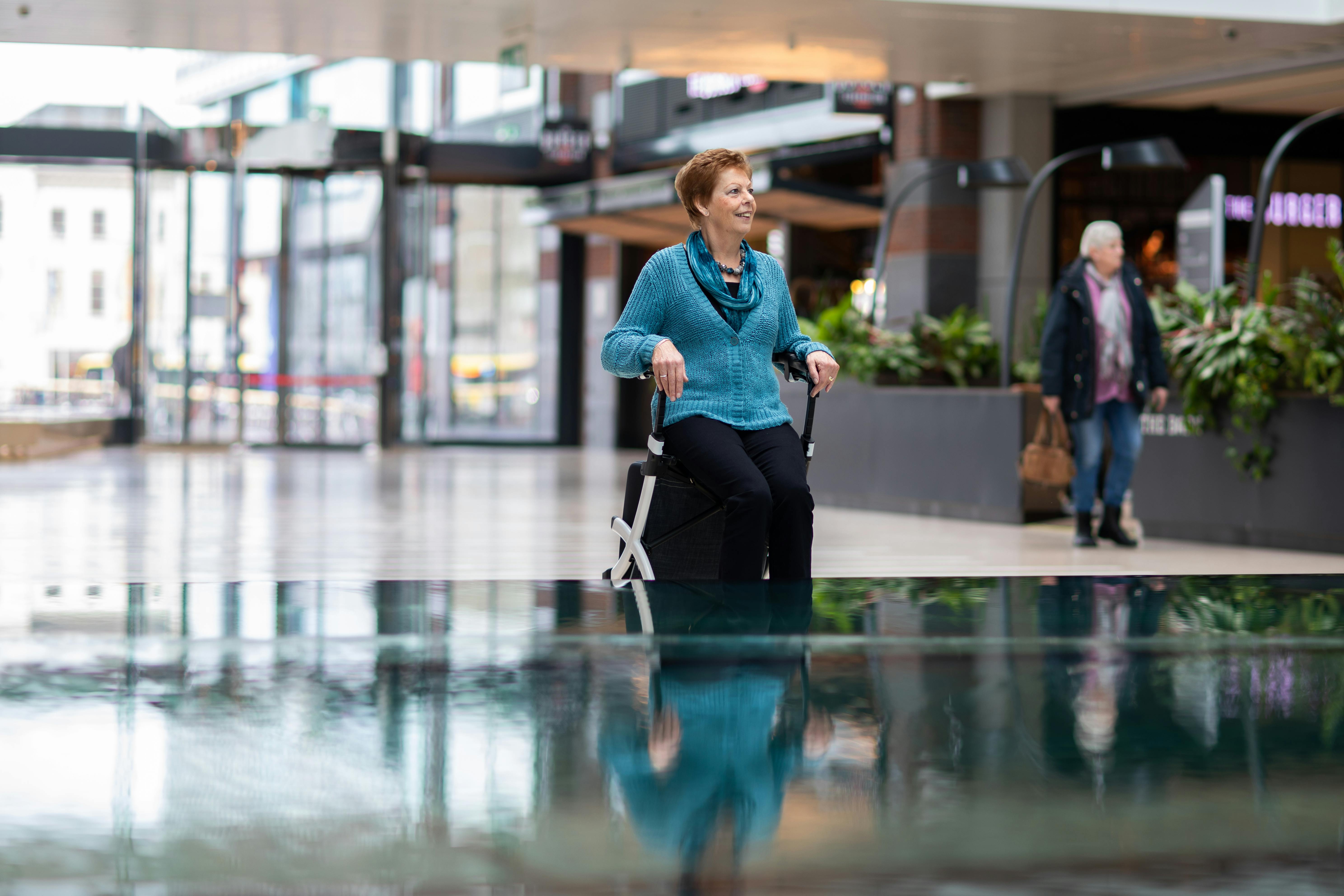 Elderly woman sitting on a walker chair in a mall, smiling thoughtfully.