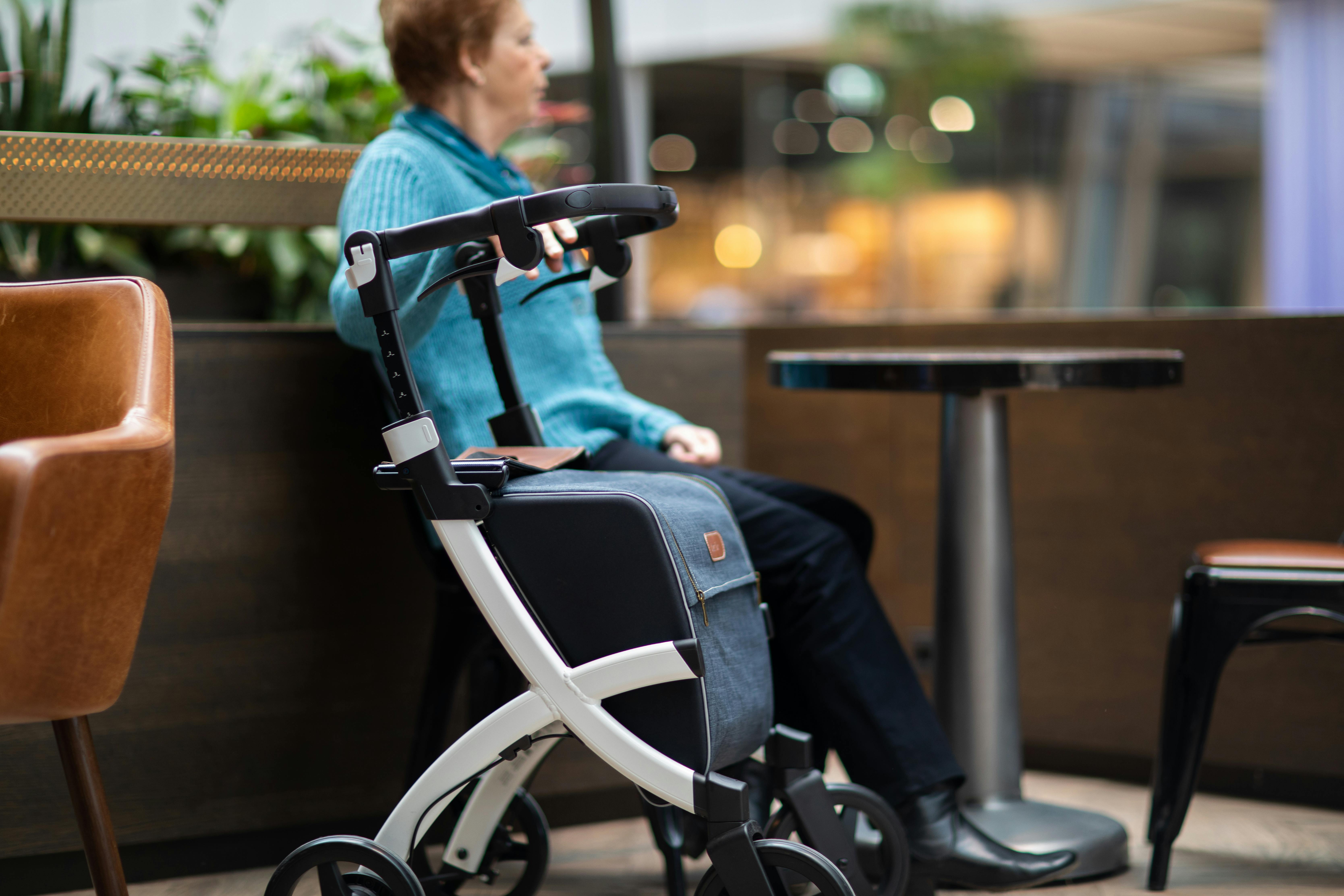 Elderly woman sits indoors in a mall with a rollator walker, enjoying leisure time.