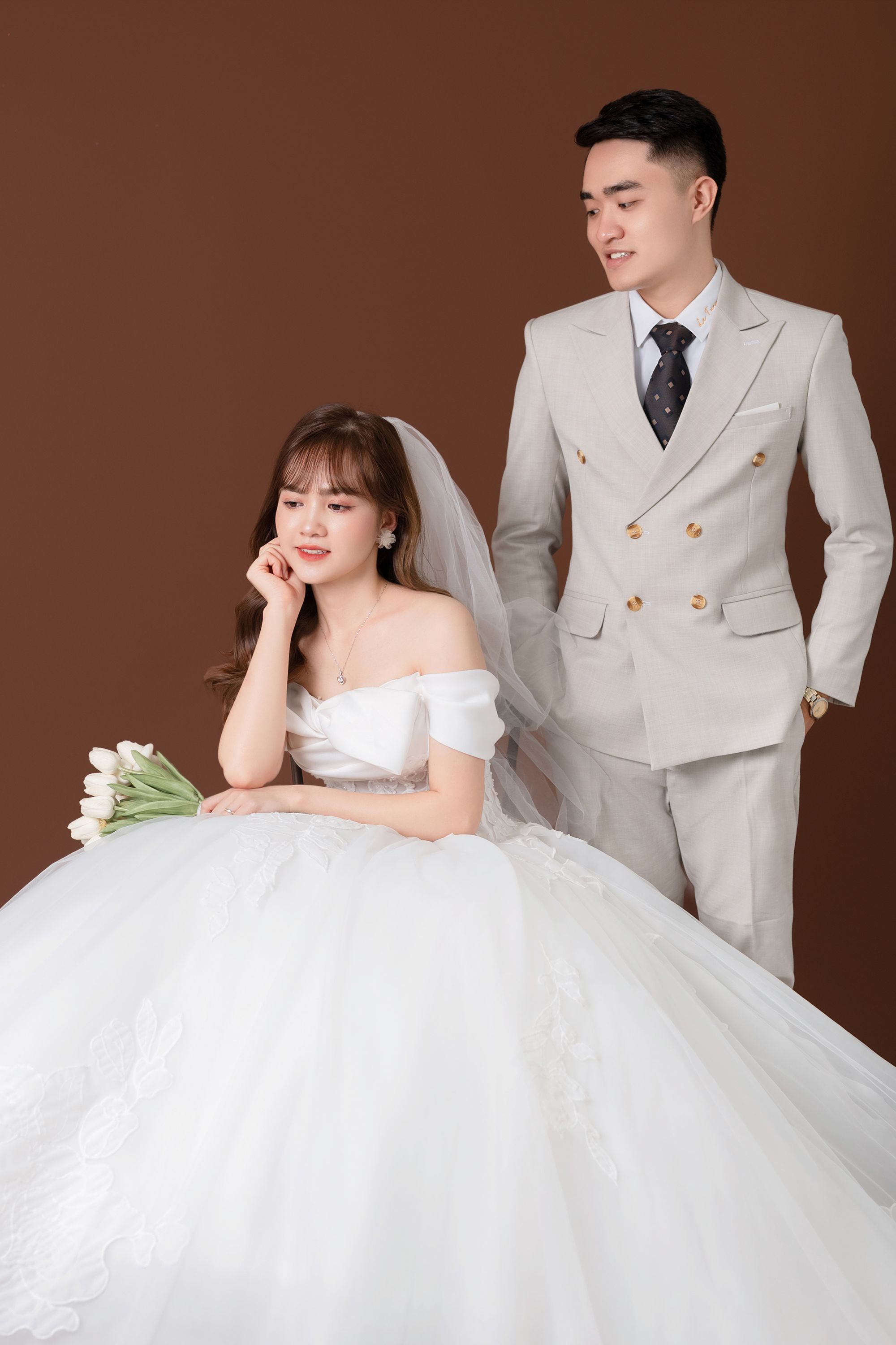 Beautiful Asian couple in stylish wedding attire captured in a studio setting, featuring a bride in white and a groom in a light suit.
