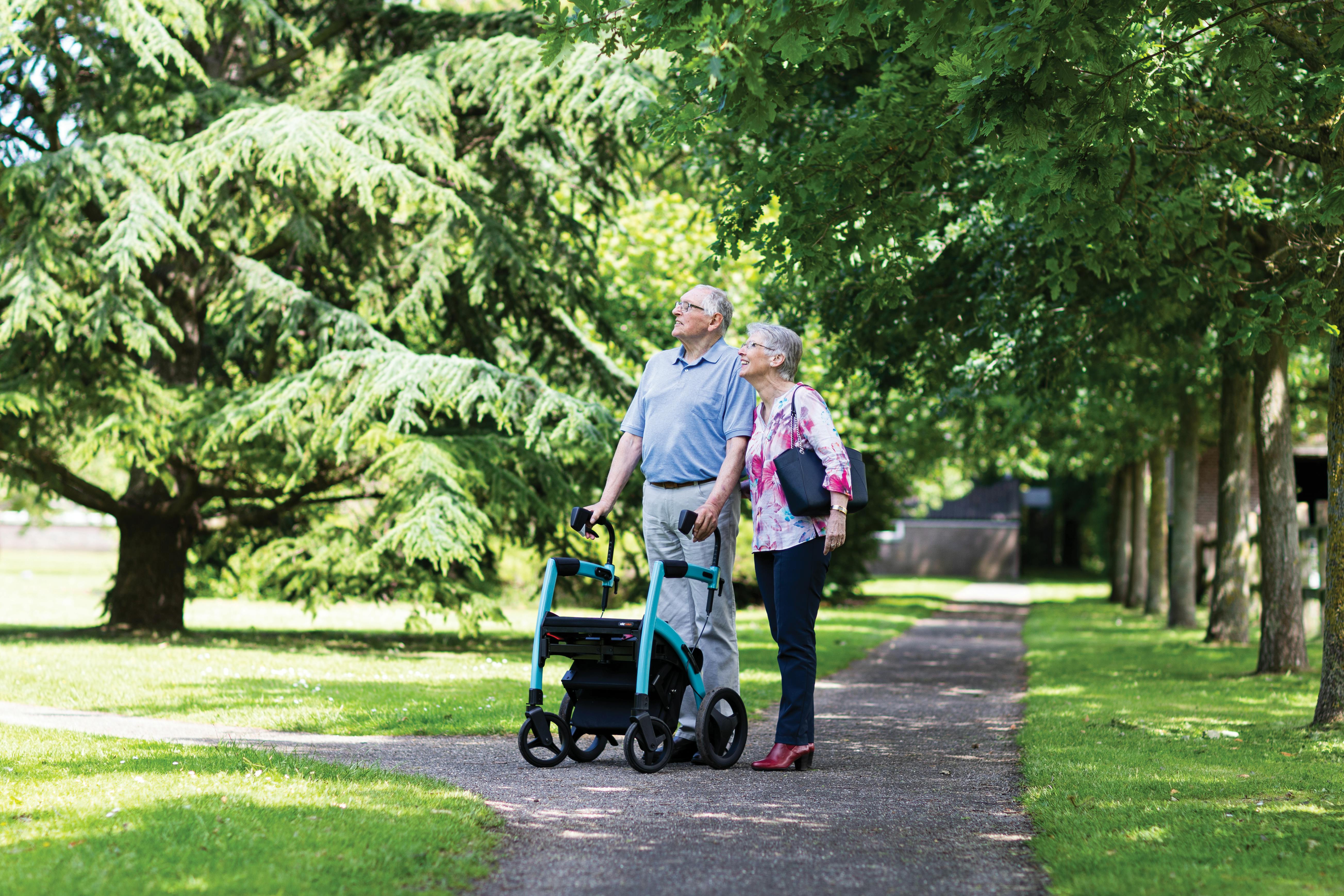 Senior couple walking with a walker in a summer park, surrounded by lush greenery.