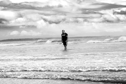 Man in T-shirt Walking on Sea Shore in Black and White