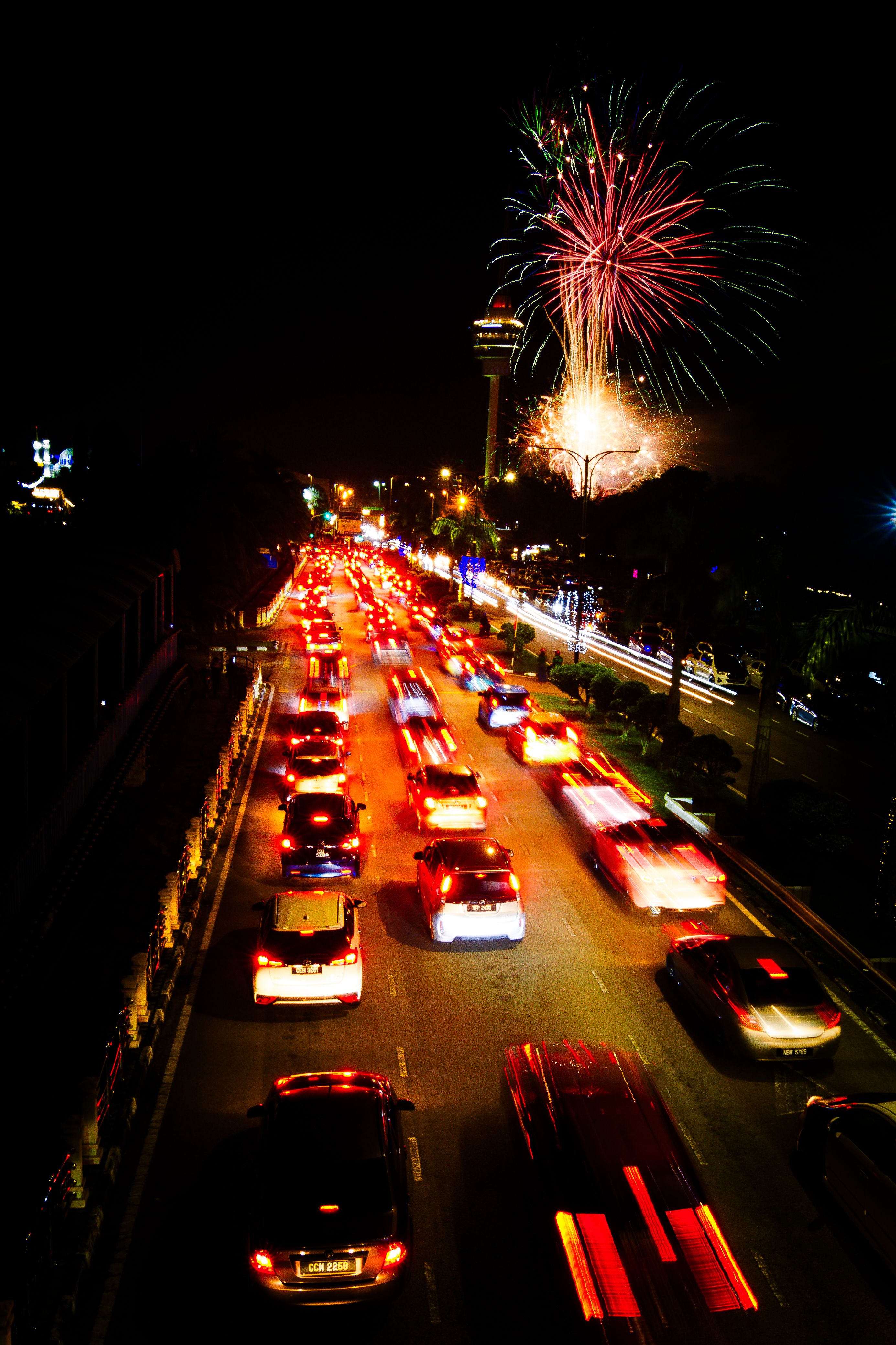 Cars drive below colorful fireworks lighting up the night sky over a busy city street.