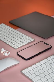 Stylish workspace featuring wireless keyboard, mouse, earbuds, smartphone, and laptop on a pink surface.