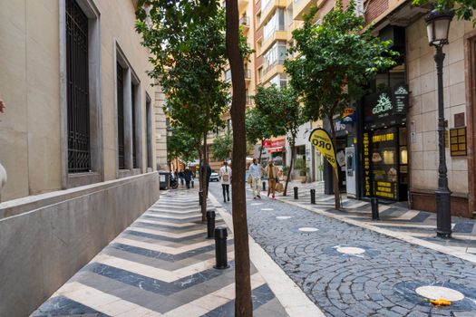 Charming urban street with cobblestones, trees, and people walking, creating a vibrant summer atmosphere.