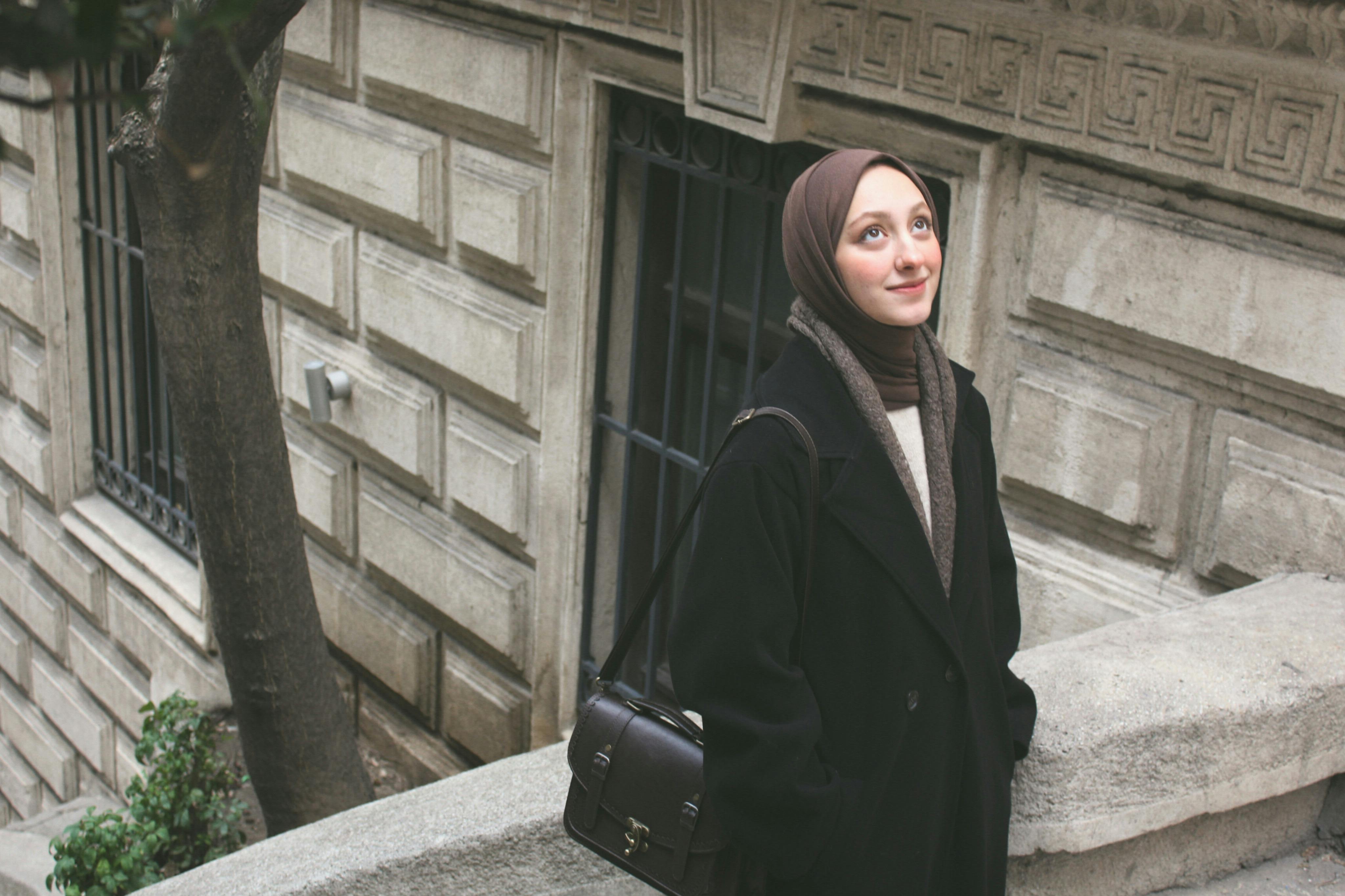 Charming young woman in a hijab with a purse amidst urban architecture, showcasing elegance and modern fashion.