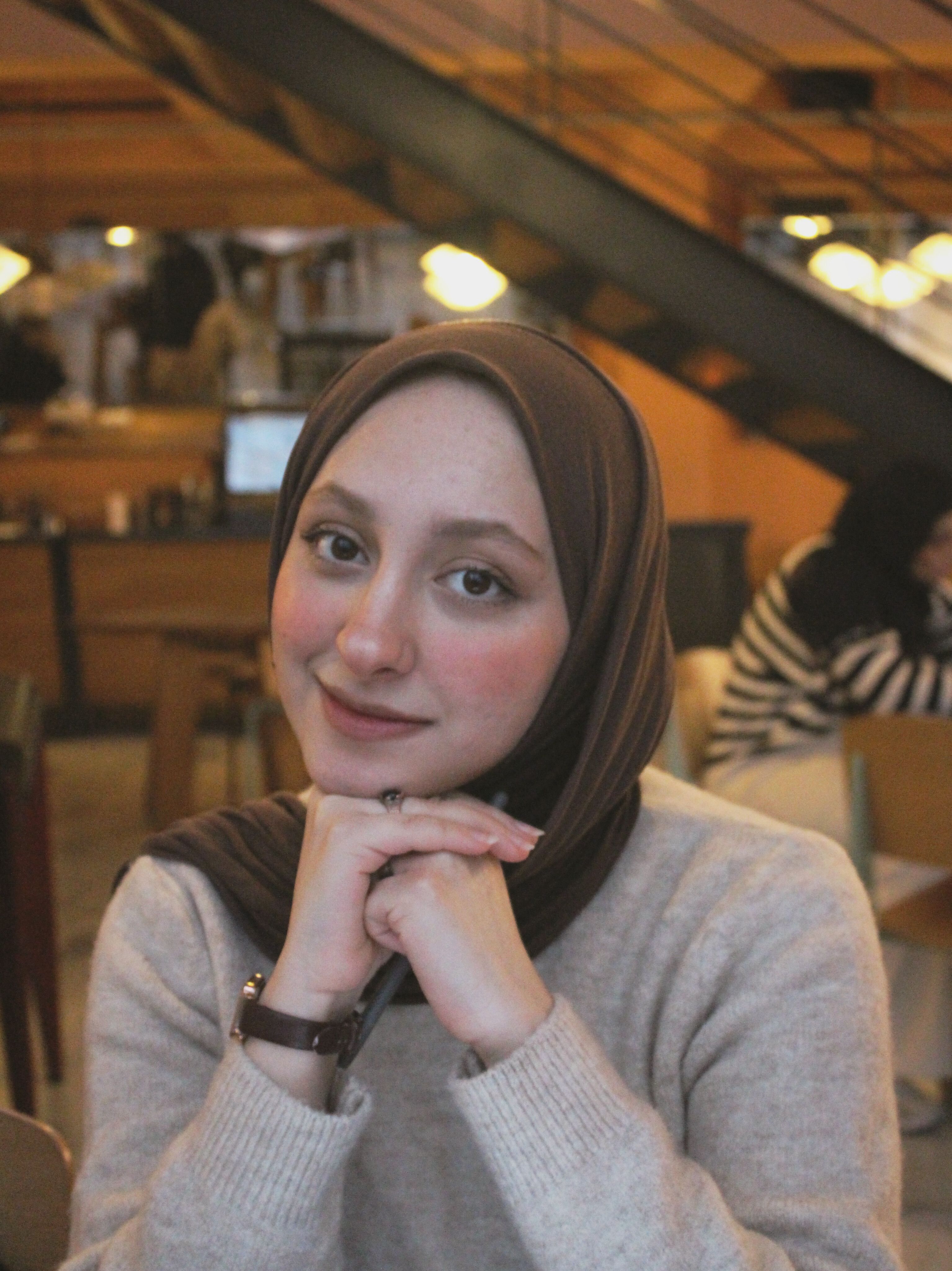 A woman in a headscarf smiling warmly in a cozy cafe environment.