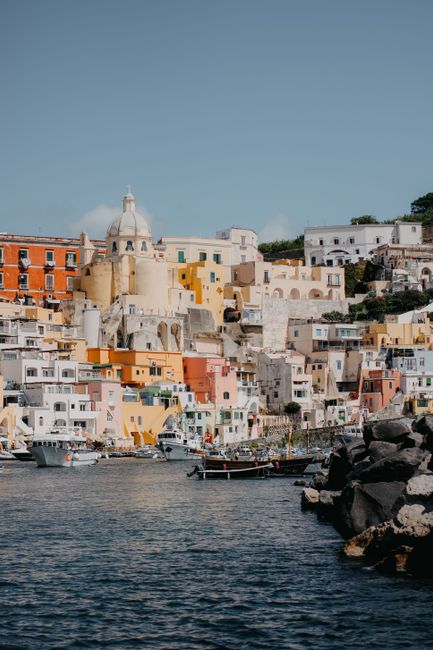Vibrant buildings line the coast of Procida, Italy, with boats docked at the shore.