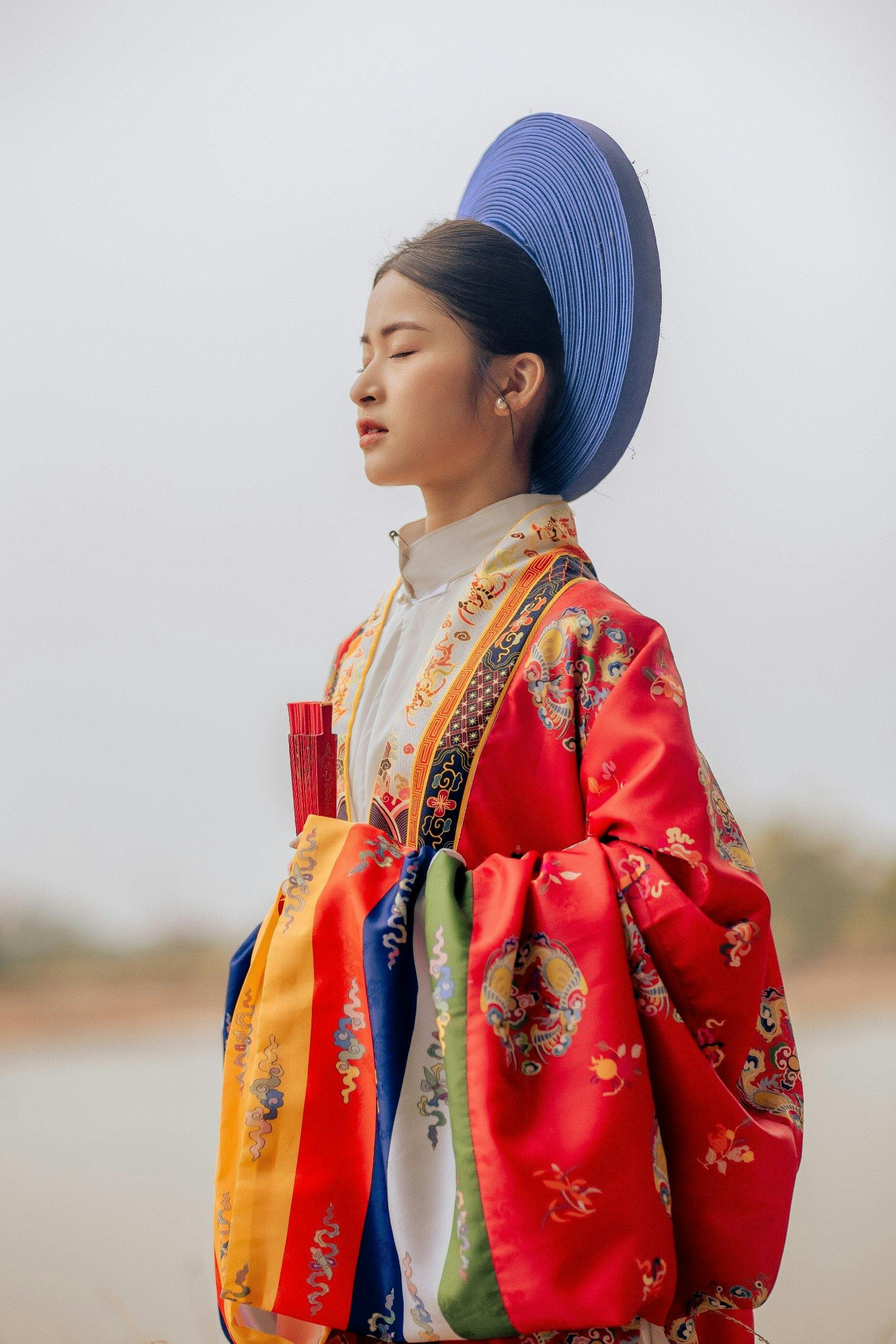 Young Vietnamese woman in traditional colorful attire, eyes closed, standing outdoors.