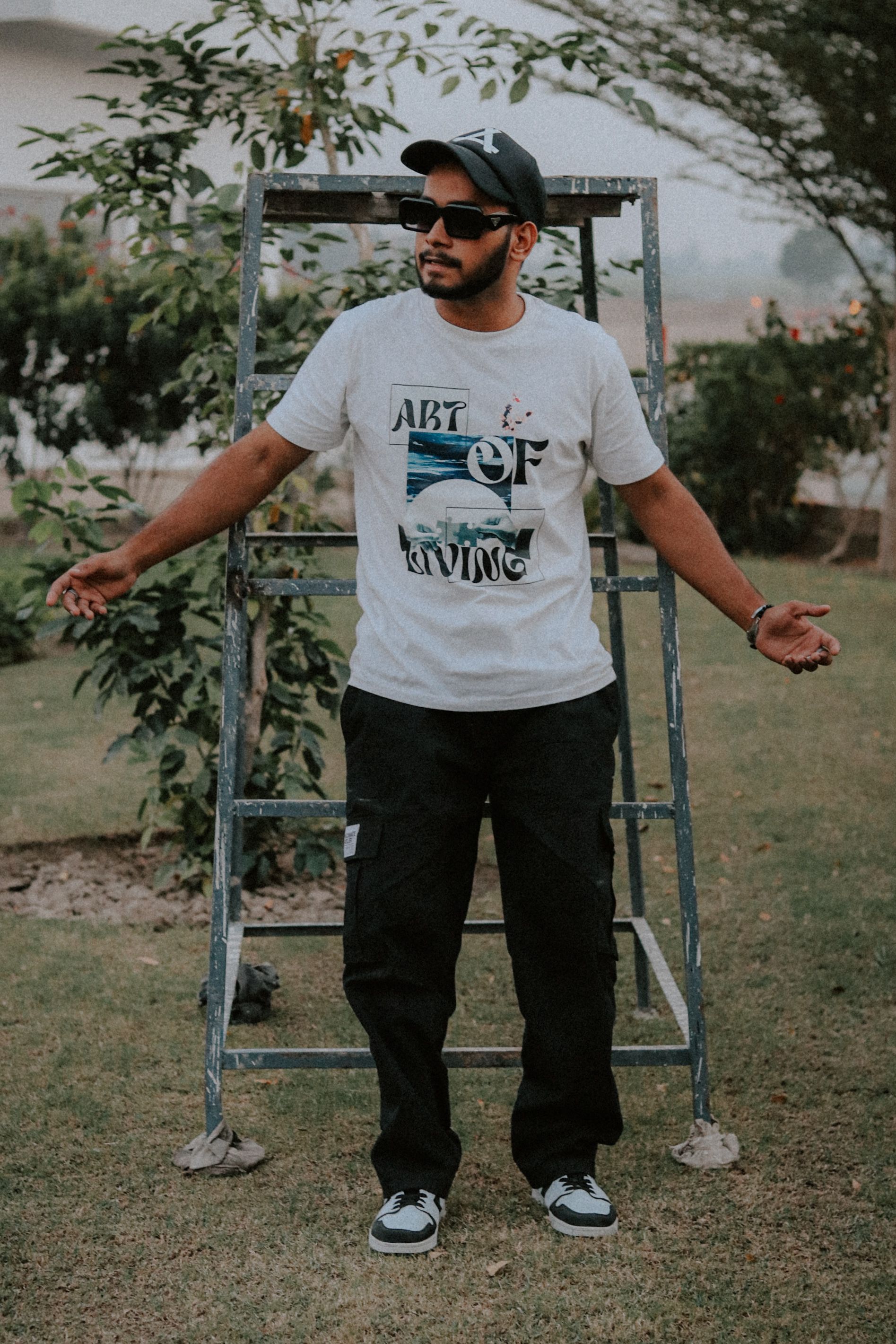 Man wearing t-shirt and cap in a yard, casual outdoor style with shades and tracksuit pants.