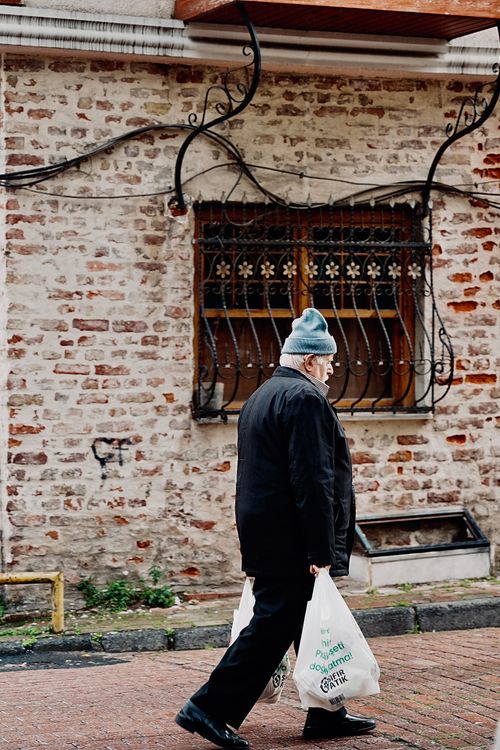 Free An elderly man walks along a brick wall carrying plastic bags, wearing a winter hat and jacket. Stock Photo