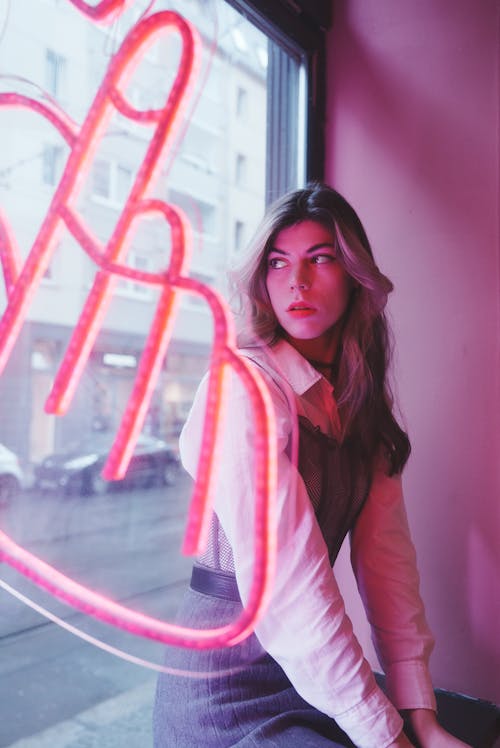 Young, Fashionable Woman Sitting by the Window with a Pink Neon Sign 