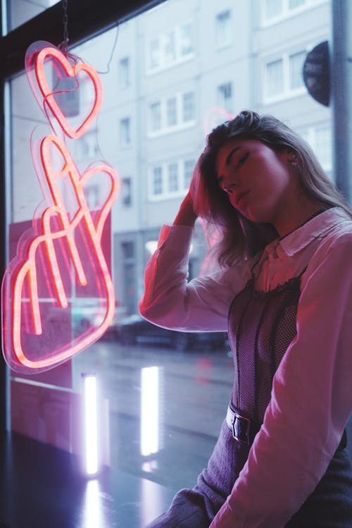 Young, Fashionable Woman Sitting by the Window with a Pink Neon Sign 