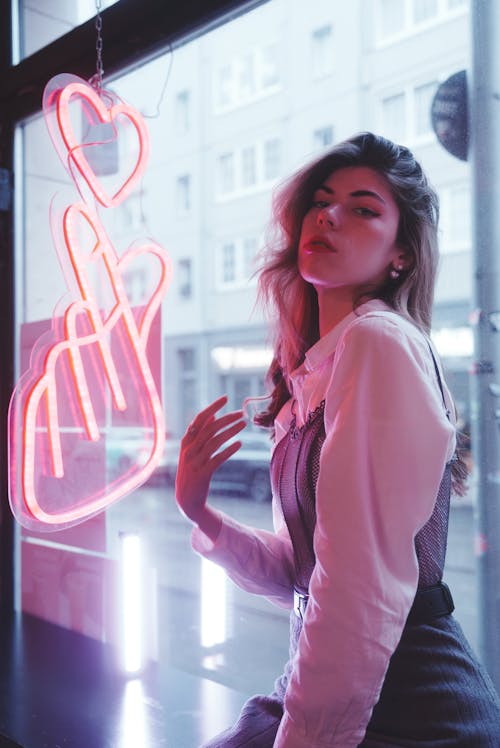 Young, Fashionable Woman Sitting by the Window with a Pink Neon Sign 