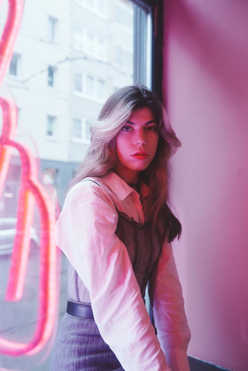 Young, Fashionable Woman Sitting by the Window with a Pink Neon Sign 