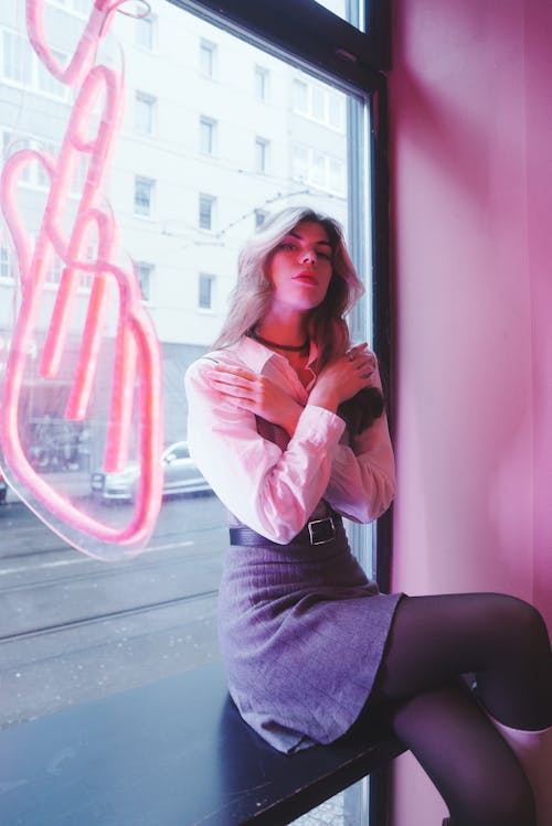 Young, Fashionable Woman Sitting by the Window with a Pink Neon Sign 