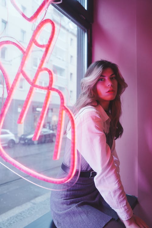 Young, Fashionable Woman Sitting by the Window with a Pink Neon Sign 