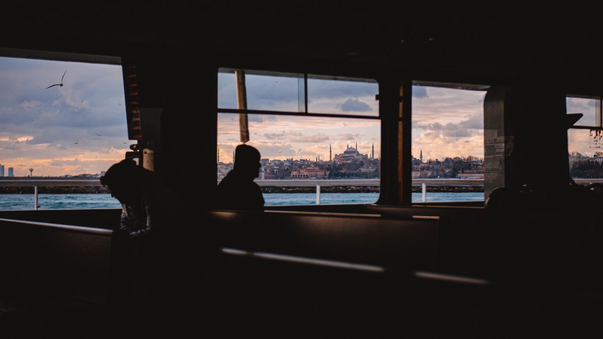 Silhouetted ferry passengers seated by the windows at dusk, a mosque skyline visible across the water