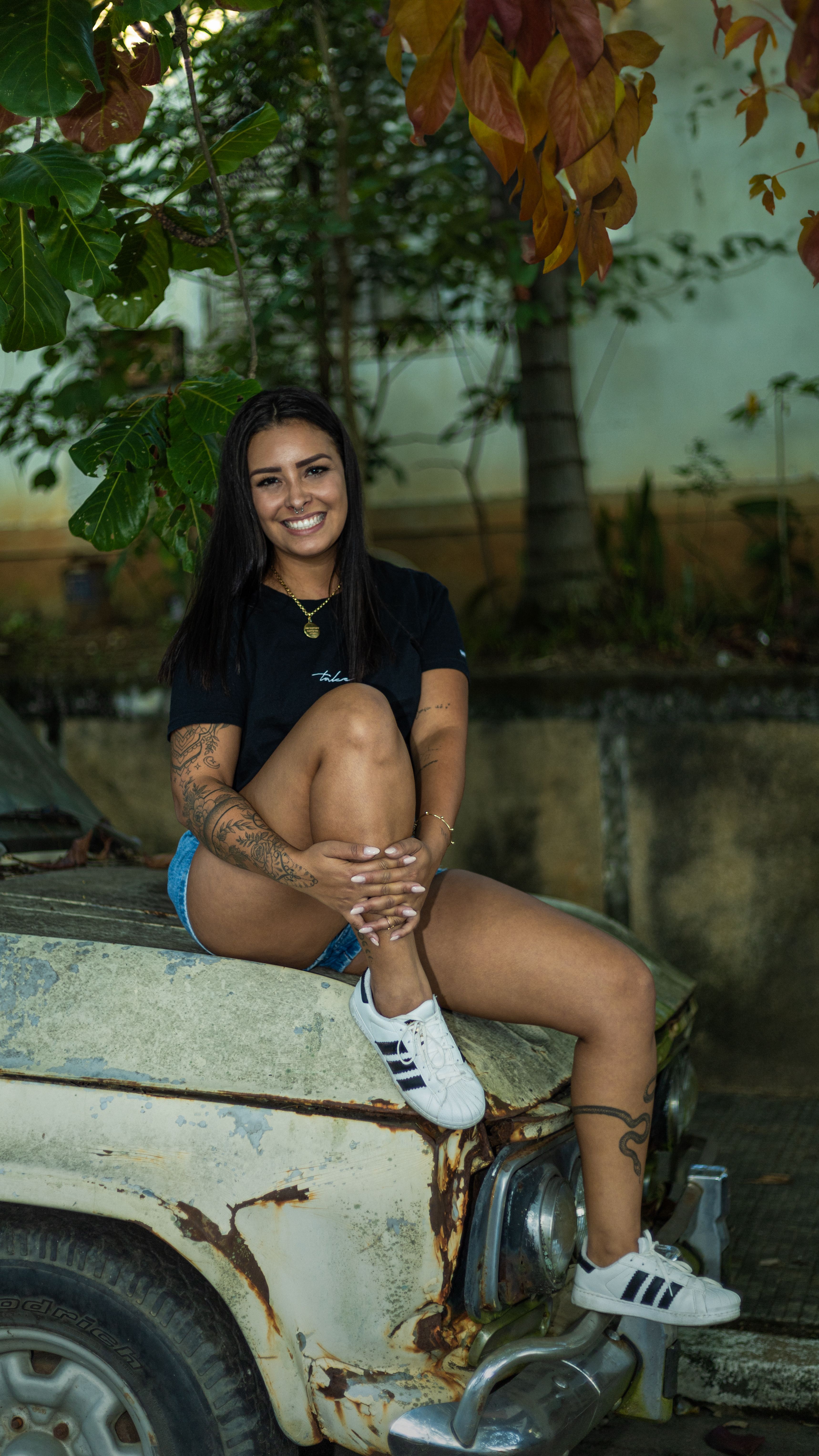 Smiling woman with tattoos sitting on an old car in an urban setting.