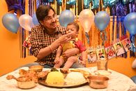 A joyful father feeds his baby son during a vibrant Indian birthday celebration with traditional food and decorations.