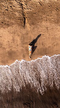 Drone captures woman in bikini relaxing on a sandy beach by the sea.