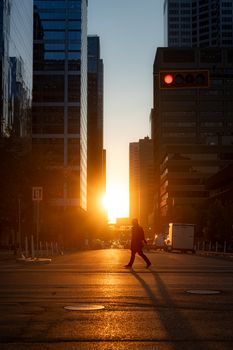 A person walking across a street in downtown Calgary, silhouetted against a stunning sunset between skyscrapers.
