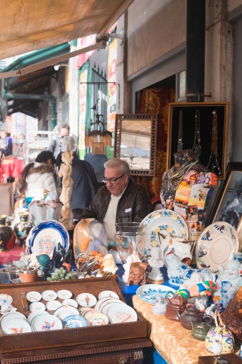 Free An elderly man sits at an antique market stall in Lisbon, Portugal, showcasing a variety of vintage items. Stock Photo
