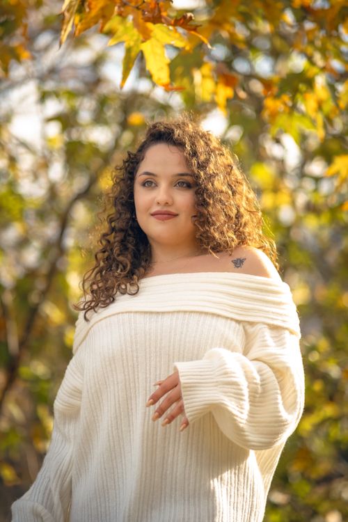 Free A woman with curly hair and a tattoo smiles in a white sweater amidst autumn leaves. Stock Photo
