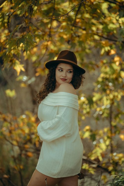 Free A fashionable woman in a white sweater and hat stands amidst autumn foliage, showcasing natural beauty and style. Stock Photo