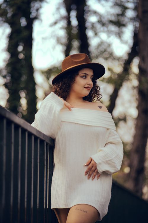 Free Elegant portrait of a woman in a white dress and hat, leaning against a railing in a wooded setting. Stock Photo