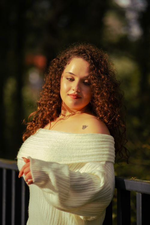 Free A woman with curly brown hair standing by a railing, wearing a white sweater. Stock Photo