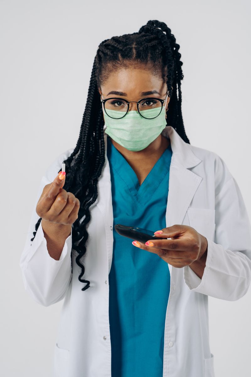 A Black woman clinician in a lab coat and scrubs holds a glucose meter and a lancet, ready to check blood sugar. Black adults face higher diabetes rates and worse complications, so early testing and the right medication matter.