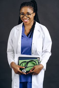 Portrait of a smiling female doctor in a lab coat holding a stethoscope, against a gray background.