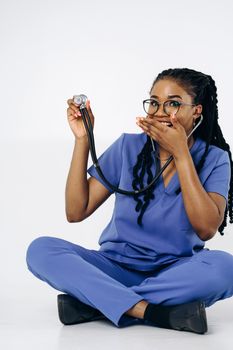 Black female doctor in blue scrubs holding a stethoscope while seated on a white background, smiling playfully.