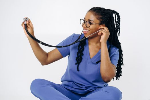 Smiling nurse in blue scrubs with braided hair holds a stethoscope against a white background.