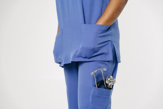 Close-up of a healthcare worker in blue scrubs with a stethoscope in the pocket, standing against a white background.