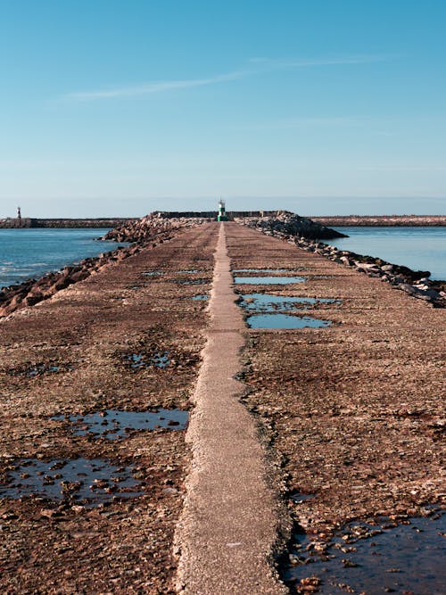 A bumpy pedestrian road with puddles along a breakwater, flanked by the ocean, leads to a small geodesic landmark, creating a symmetrical diminishing perspective. Praia do Cabedelo, Figuei...
