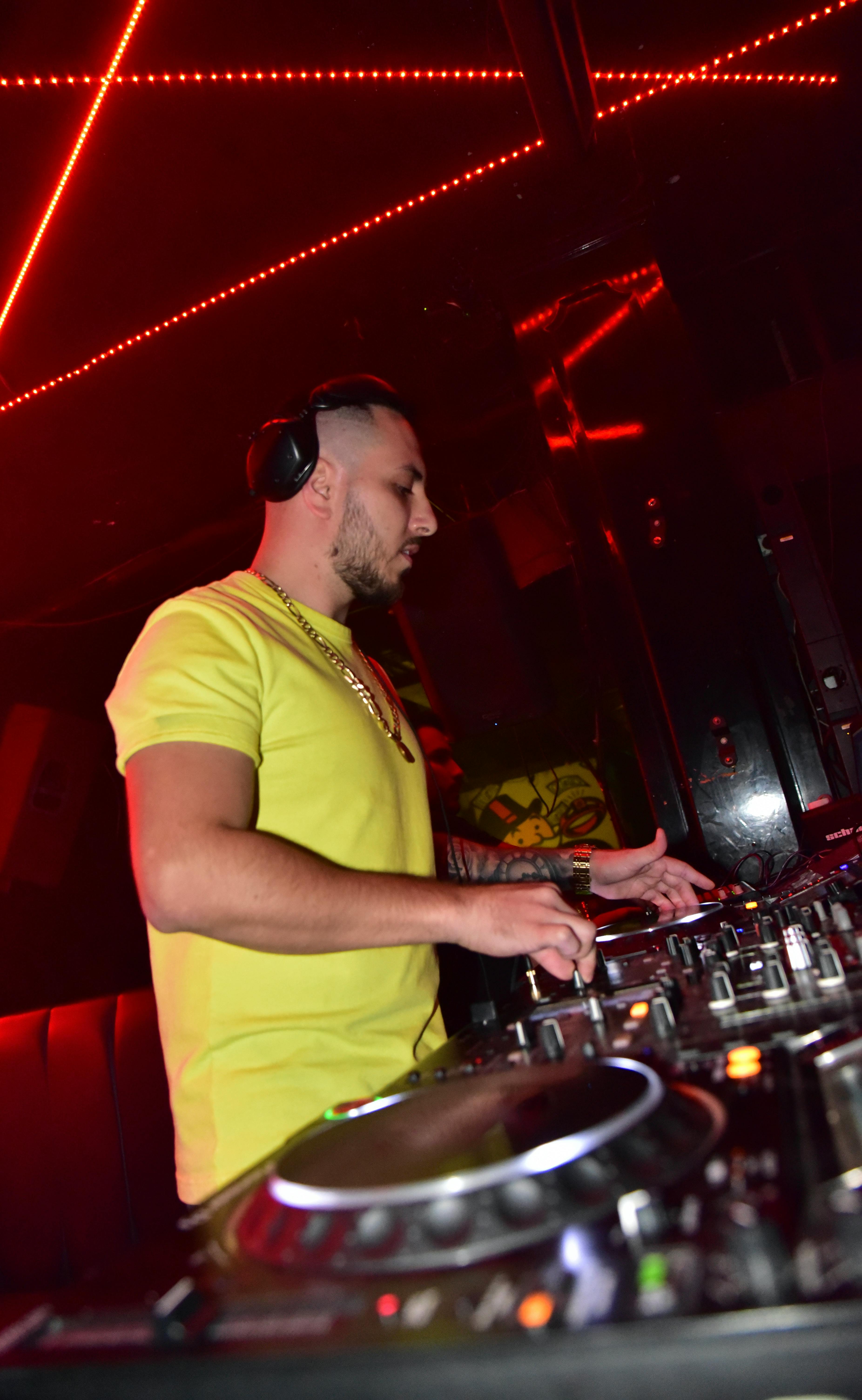 A DJ in a yellow t-shirt spins tracks in a neon-lit nightclub, creating a lively atmosphere.