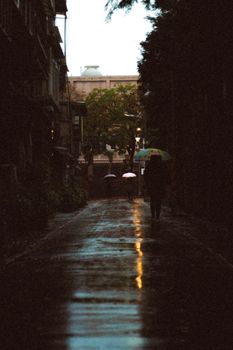 A dimly lit urban alley at dusk with people walking under umbrellas, reflecting a rainy mood.