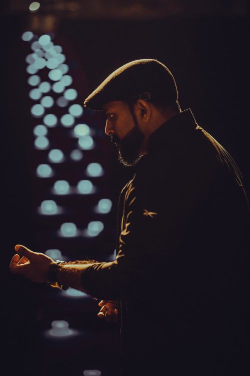 Free Silhouette of a man with a beard wearing an ivy cap, standing against illuminated lights creating a bokeh effect. Stock Photo