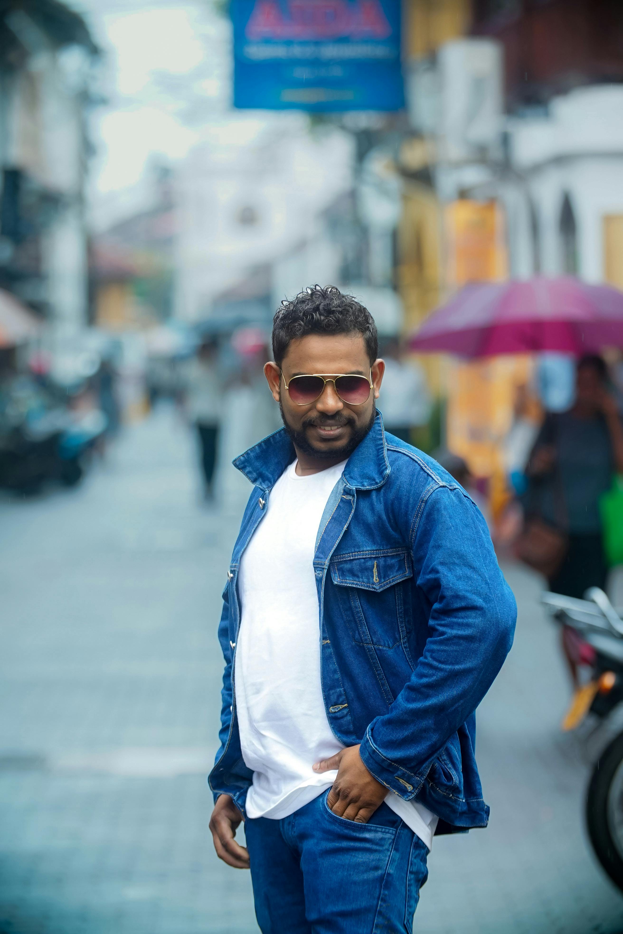 Stylish man in denim jacket poses confidently on an urban street with blurred city background.