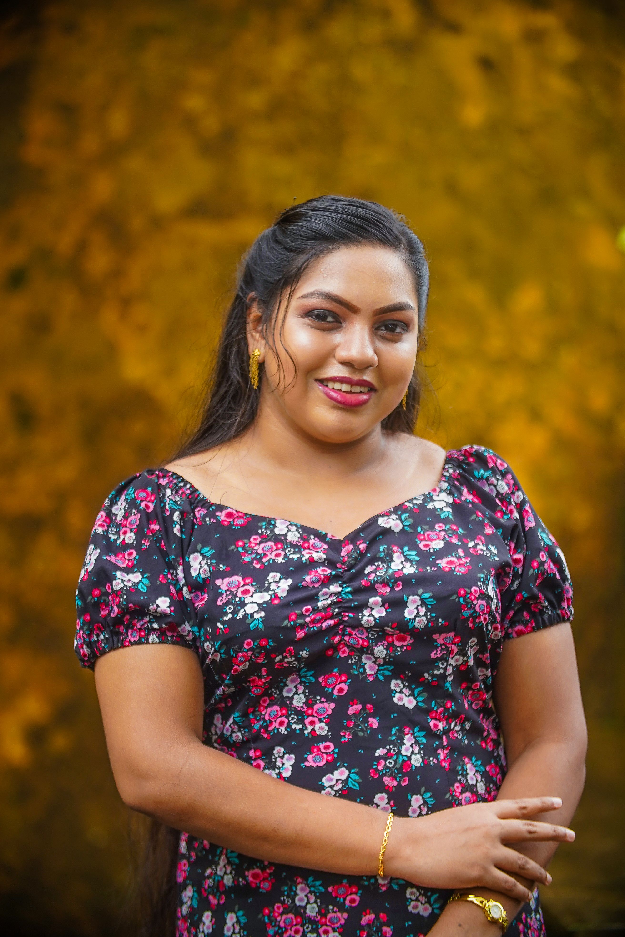 Portrait of a smiling woman in a floral dress standing against a warm, autumn-colored background.