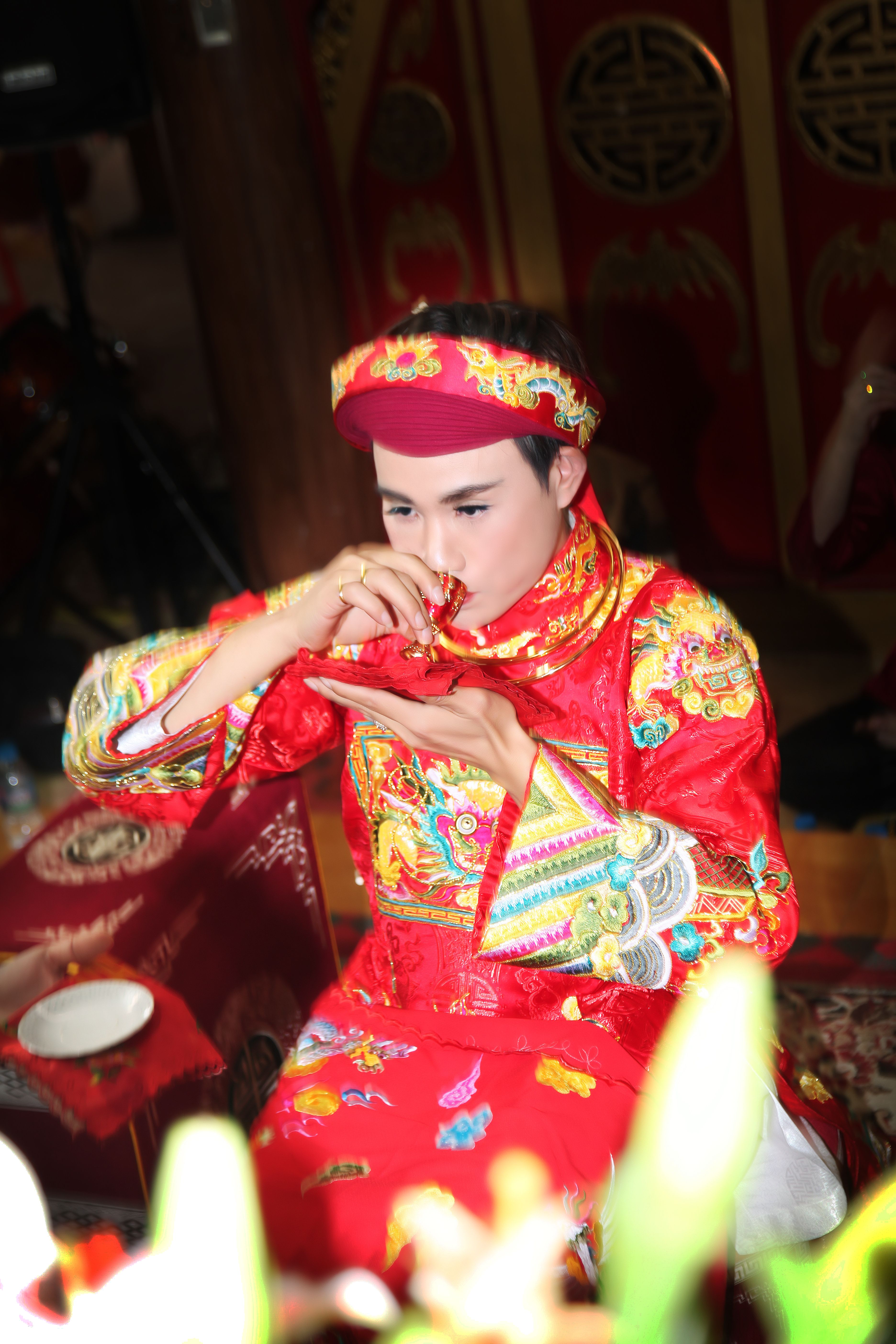 An adult in traditional Asian clothing drinks ceremonially. Captured indoors with vibrant colors.