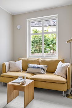 Bright living room featuring a cozy yellow sofa, pillows, lamp, and wooden table.
