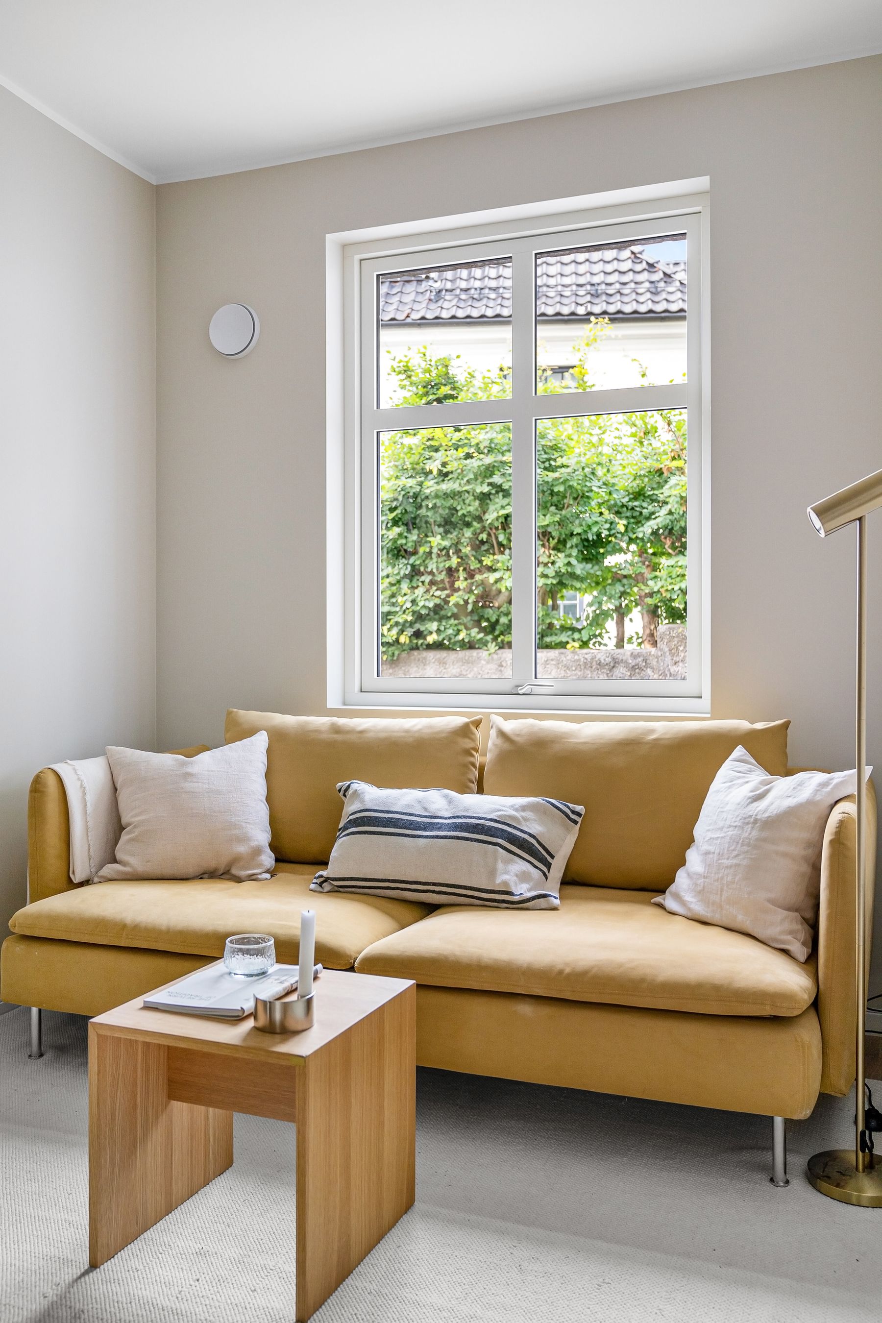 Mustard yellow sofa beneath a large window in a calm neutral living room with pale walls and a light wood table