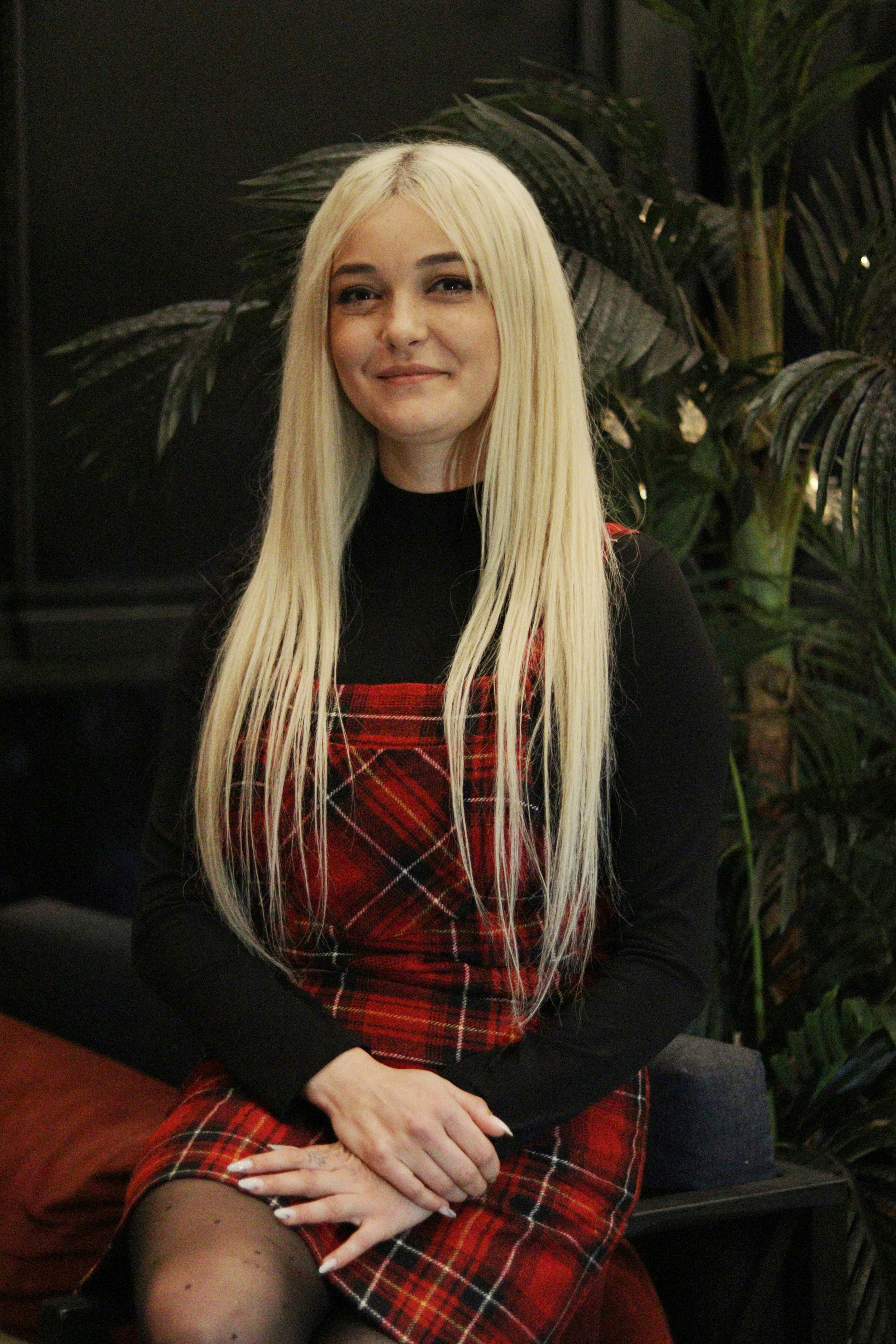 Blonde woman in a red tartan dress, sitting and smiling indoors, fashion portrait.