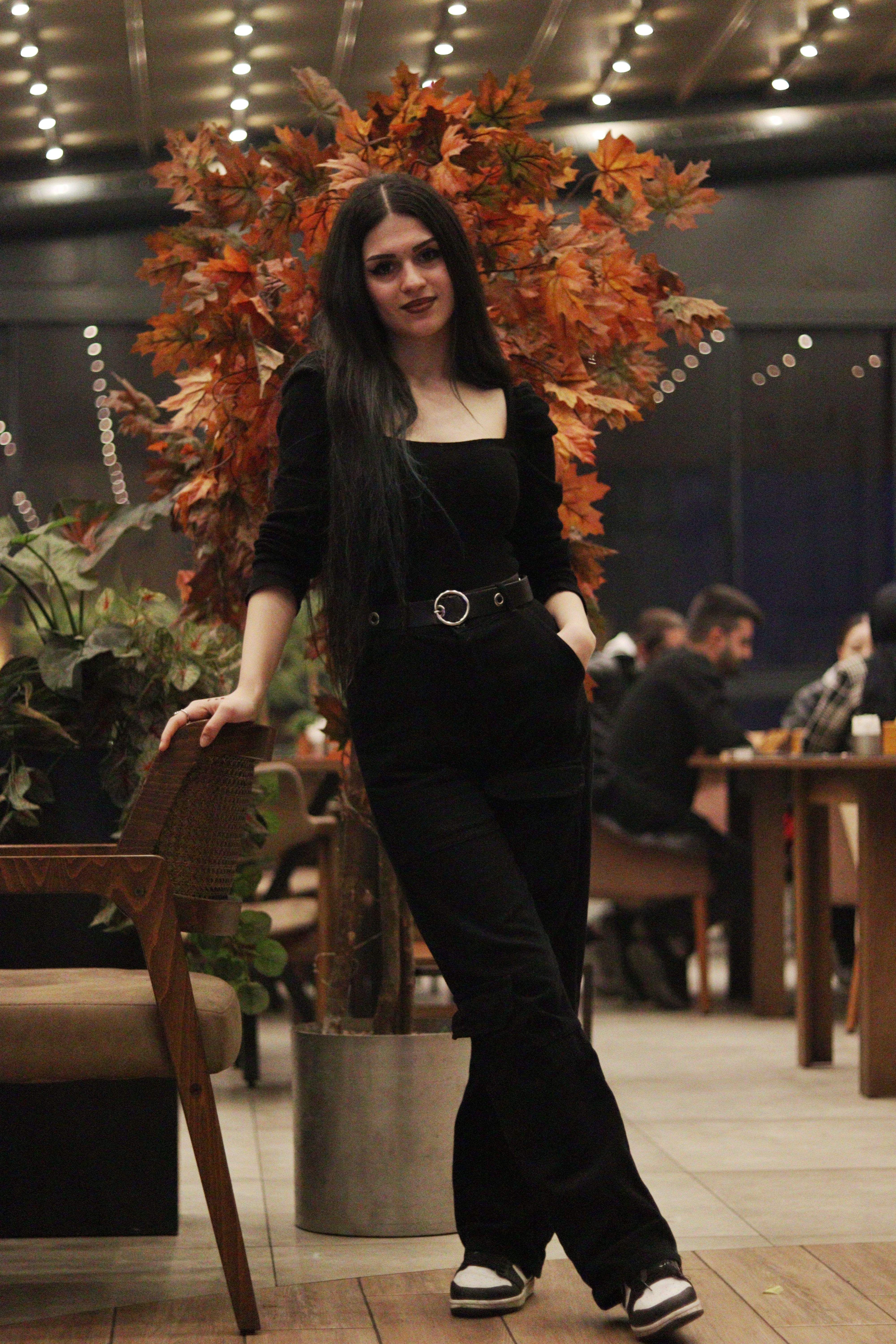 A stylish woman in a black outfit standing in a cozy cafe with autumn leaves decor.